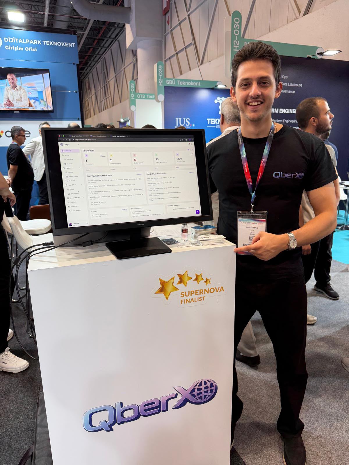 Berker Yenal, GITEX AI Türkiye 2026 Hall 2'deki QBERX standında; ekranda QRegu uyum panosu, pano üzerinde Supernova Finalist rozeti